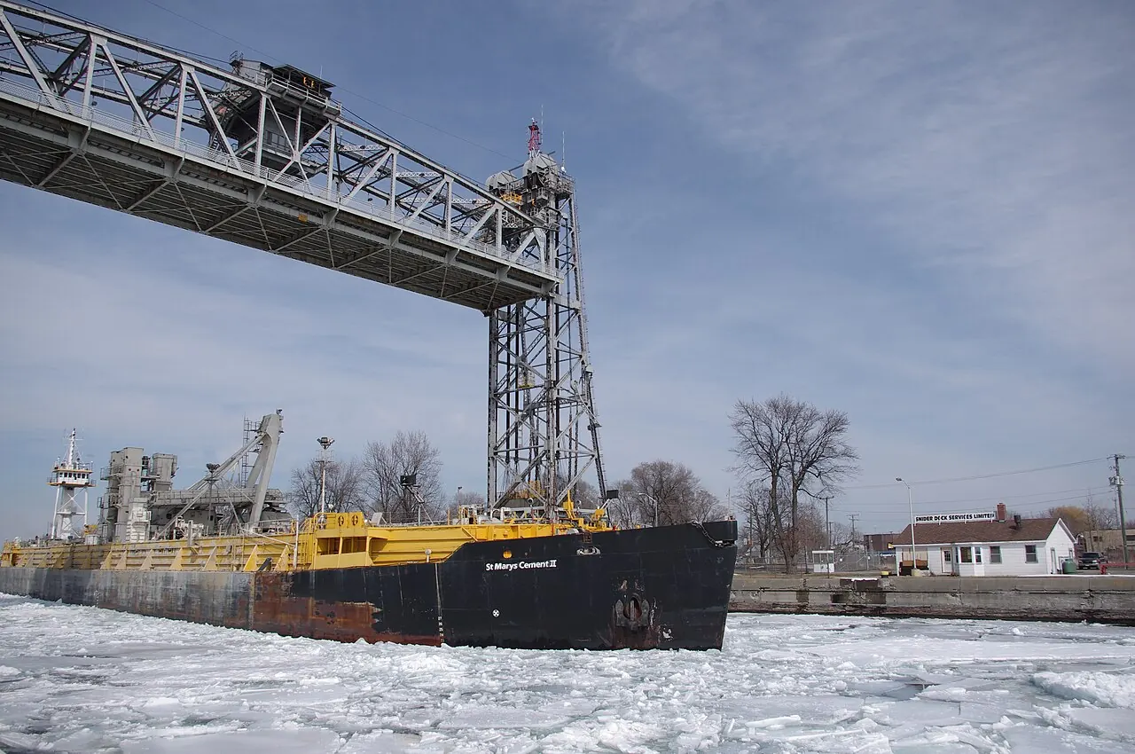A lake freighter passing under the raised span of a lift bridge on the Welland Canal at Port Colborne, with ice along the canal wall.
